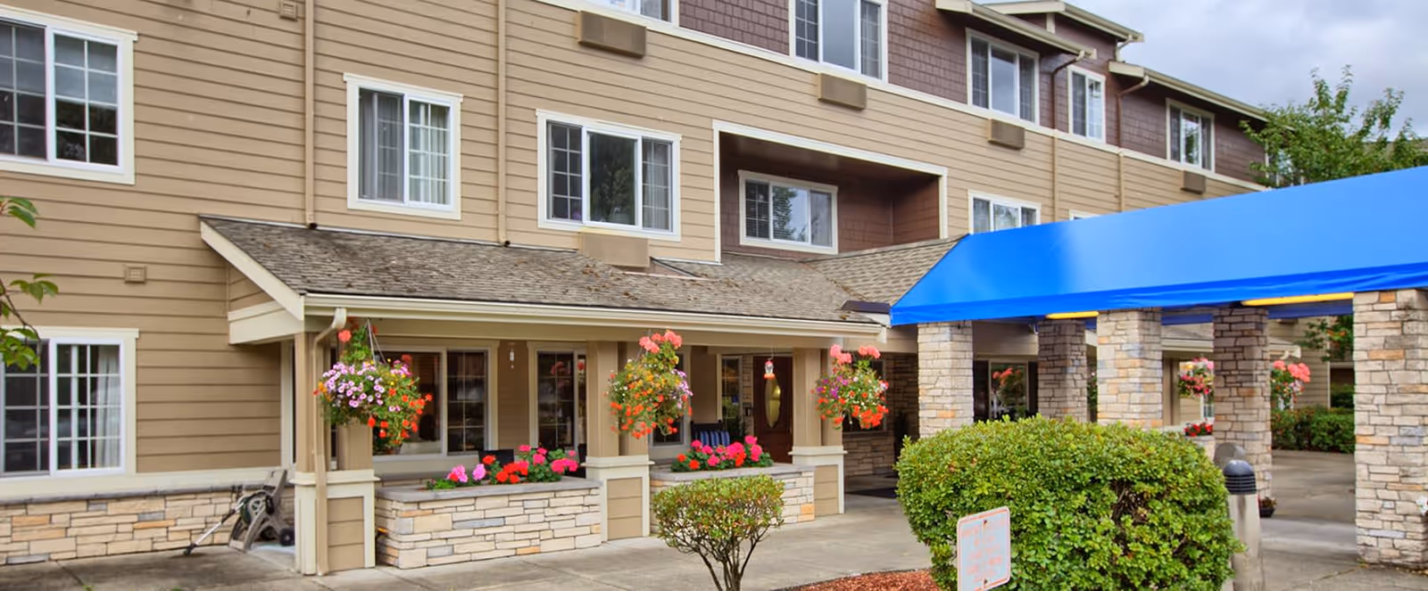 Exterior view of a senior living facility with beige siding and stone accents. There are hanging flower baskets with pink and red flowers along the covered entrance area, which has a blue canopy supported by stone pillars. Bushes and small trees are planted around the entrance.