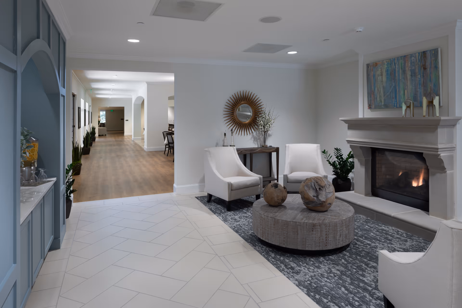 A cozy living room area in The Blake at Hollingsworth Park featuring a lit fireplace with a mantel decorated with artwork and small sculptures. The room has three white upholstered chairs arranged around a round wooden coffee table with decorative spheres. A sunburst mirror and a small table with a vase of flowers are against the wall. The floor transitions from light-colored tile to a patterned rug, and a hallway with wood flooring and potted plants extends into the background.