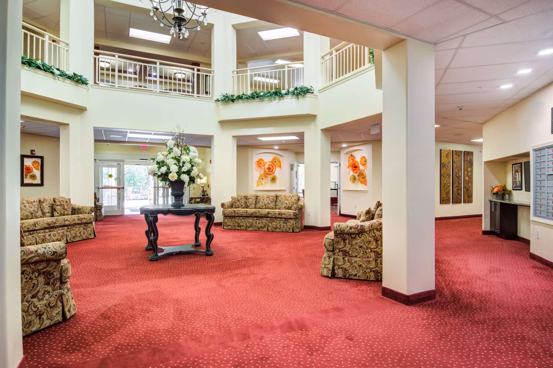 Spacious senior living facility common area with red carpet flooring, patterned sofas arranged around a central black table holding a large floral arrangement. The room features high ceilings with a chandelier, cream-colored walls, decorative artwork, and an upper balcony railing overlooking the space.