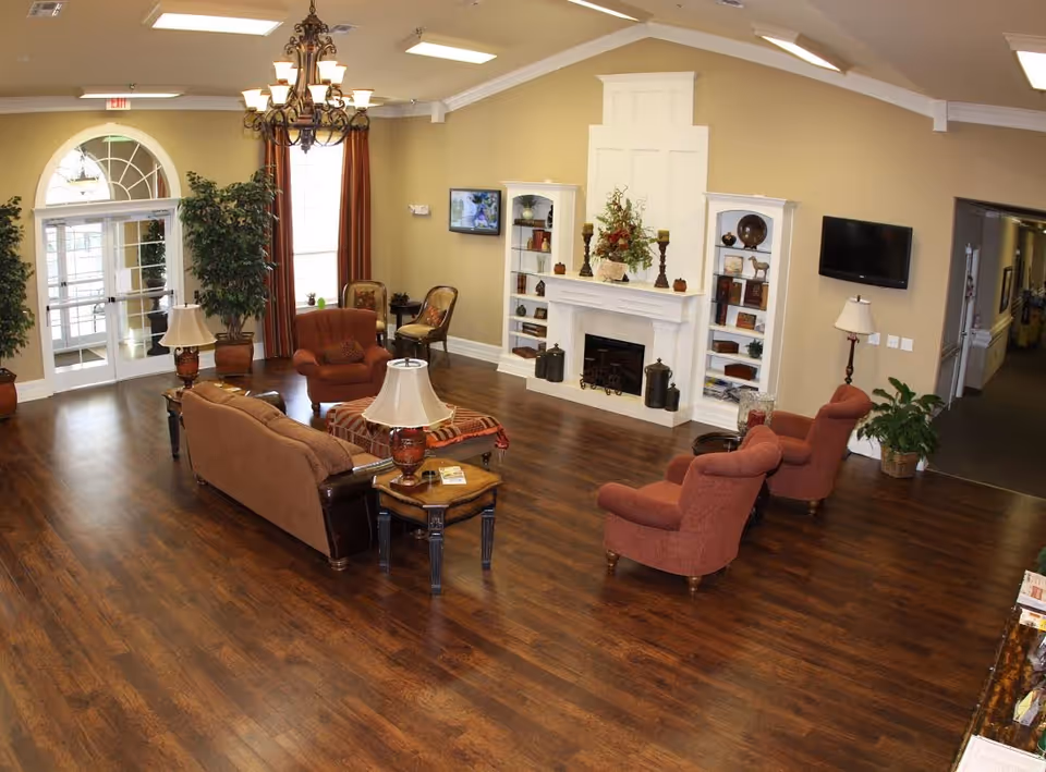 A spacious senior living facility common area with wooden flooring, beige walls, and a white fireplace flanked by built-in shelves. The room is furnished with several upholstered chairs and sofas in warm tones, a chandelier hanging from the ceiling, potted plants, and two wall-mounted flat-screen TVs. Large windows with red curtains and glass double doors allow natural light into the room.