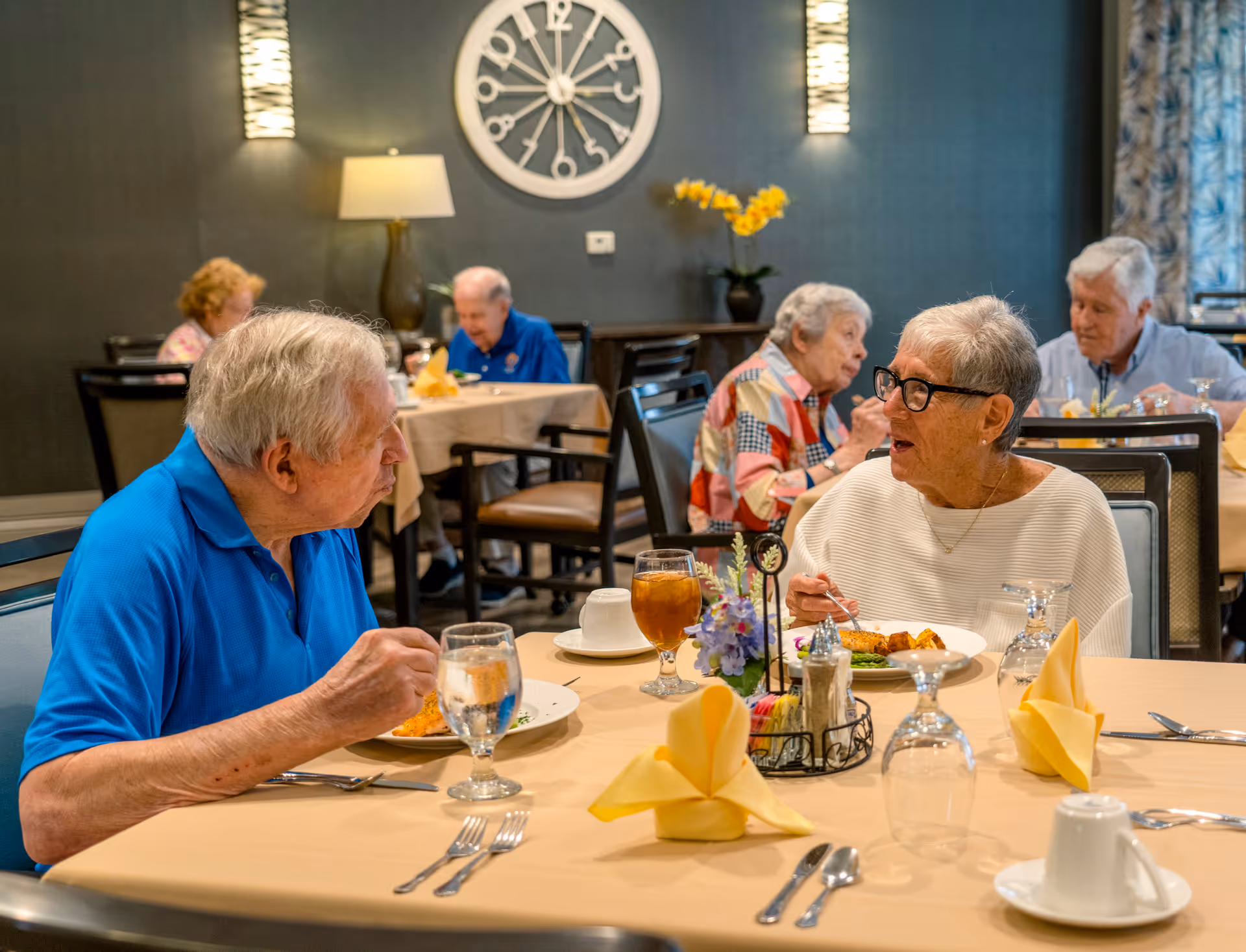 Several elderly people seated at tables in a dining room, eating and conversing. The room has a large decorative clock on the wall, wall sconces, and a lamp. Tables are set with yellow folded napkins, glasses, and plates of food.