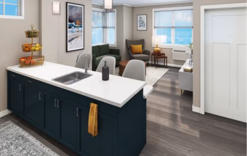 A modern open-concept living space featuring a kitchen island with a sink and soap dispenser, dark blue cabinets, and a yellow towel hanging on the side. In the background, there is a cozy living room area with a green sofa, a gray armchair with a yellow pillow, a small wooden coffee table, a side table with a lamp, and large windows letting in natural light.