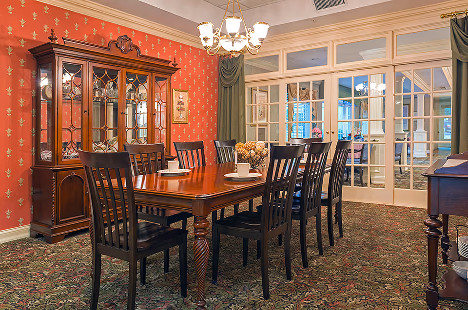 A traditional dining room with a long wooden dining table surrounded by eight dark wooden chairs. The table is set with white cups and saucers and a decorative centerpiece. Behind the table is a large wooden china cabinet with glass doors. The walls are covered in red wallpaper with a gold fleur-de-lis pattern, and the floor is carpeted with a floral design. French doors with glass panes lead to another room, and a chandelier hangs from the ceiling.