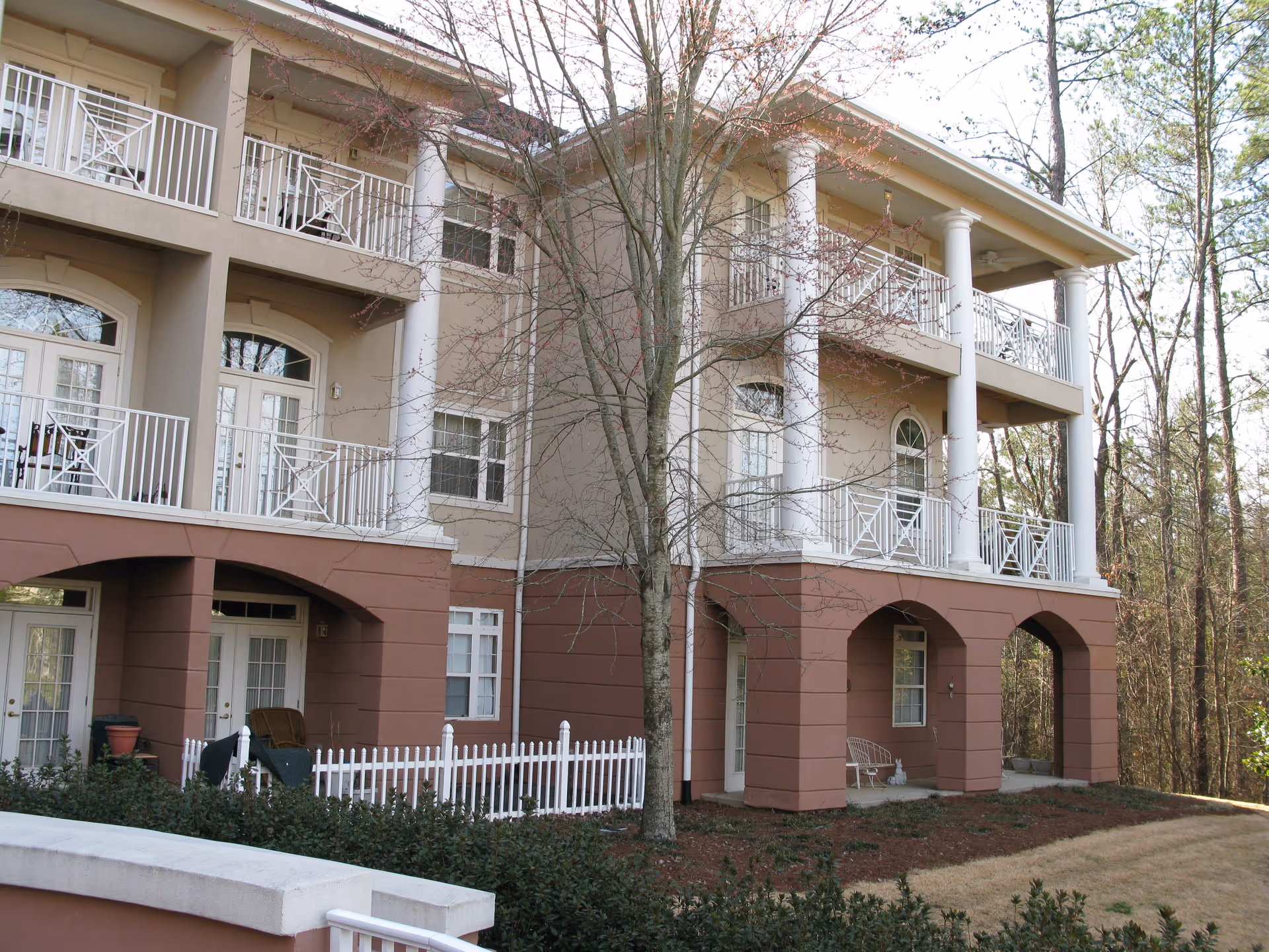 Exterior view of a multi-story residential building with beige and brown walls, white railings on balconies, large white columns, and a small fenced garden area in front. Trees with sparse branches are visible around the building.