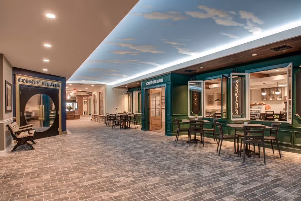 Indoor hallway area designed to resemble a small town street with a sky-painted ceiling. On the left is an entrance labeled 'County Theater' with double doors and benches nearby. On the right, there is a green storefront labeled 'Cafe on Main' with tables and chairs outside and inside seating visible through open windows.