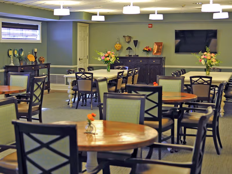 Dining/activity room with multiple round and rectangular tables and chairs, floral centerpieces, and a wall-mounted TV.