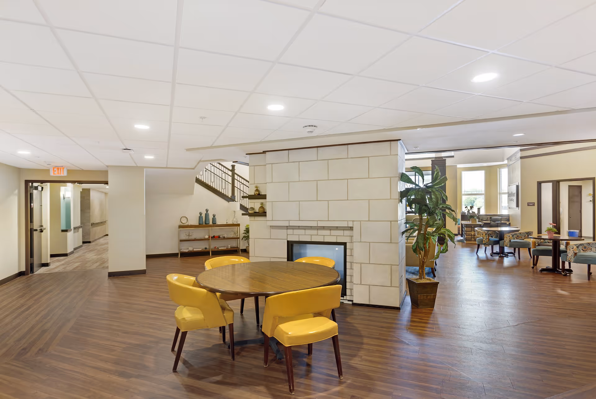 A spacious senior living common area with a round wooden table surrounded by four yellow chairs in the foreground. The room features a central stone fireplace, wood flooring, and a potted plant beside the fireplace. In the background, there are additional seating areas with tables and chairs, large windows letting in natural light, and a staircase leading upstairs.