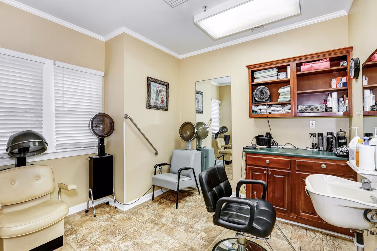 Interior of a hair salon room with beige walls and tiled floor. The room contains salon chairs, hair dryers, a large mirror, a sink, and wooden cabinets stocked with towels and hair care products. There is a framed picture on the wall and a window with white blinds.