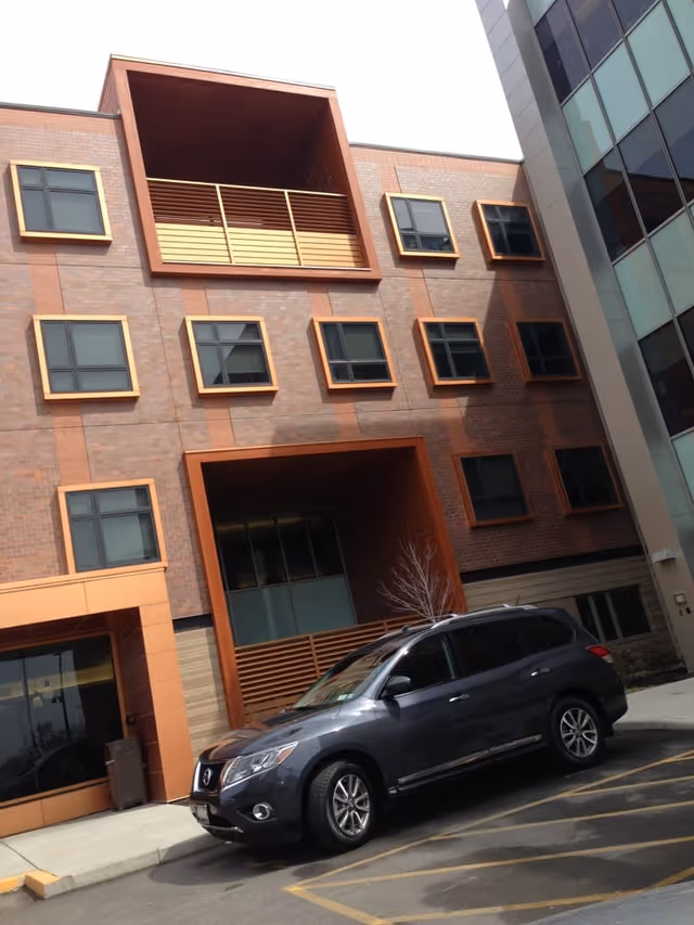 Exterior front facade of a multi-story brick building with square windows and a dark SUV parked in front.