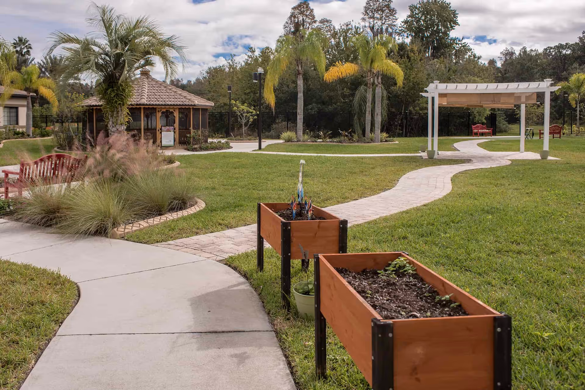 Landscaped outdoor courtyard with winding paved paths, raised wooden planters, palm trees, a gazebo and a pergola with seating.