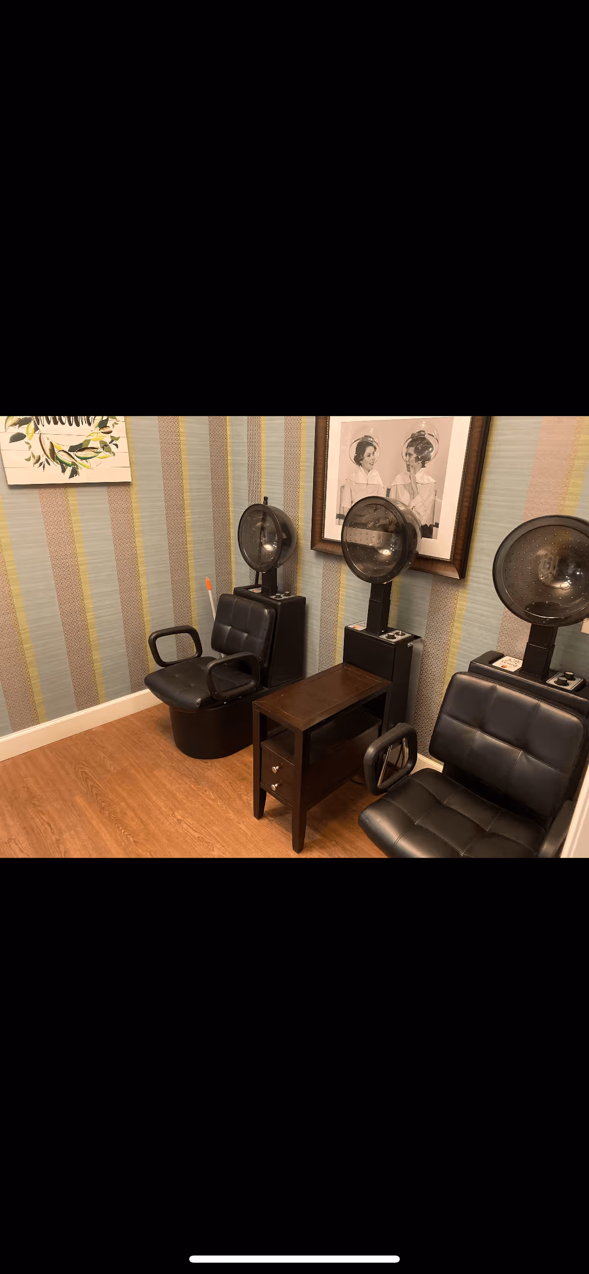 A small salon area with two black vintage hair drying chairs and hair dryers mounted on stands. The room has striped wallpaper in muted tones and a wooden floor. There is a small wooden table with drawers between the chairs. A framed black and white photo of two women with vintage hair dryers is hung on the wall.