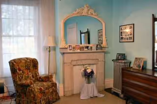 Cozy living room with a decorative fireplace, mirror, floral armchair, and dresser against turquoise walls.