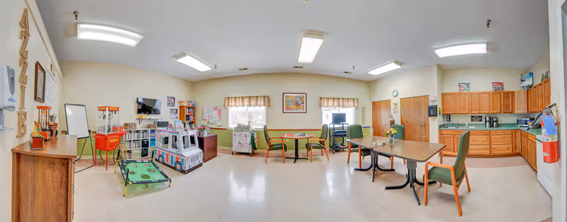 Wide, well-lit interior activity and dining room with tables, chairs, a kitchenette, and a small play/activity area including a toy car.