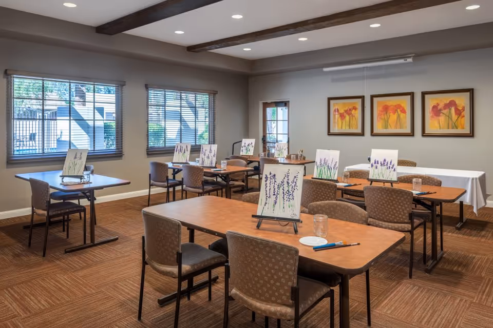 A well-lit activity room with several tables and chairs arranged for a painting session. Each table has a canvas on an easel displaying a lavender painting, along with paintbrushes, water jars, and palettes. The room has large windows with blinds, three framed floral paintings on the wall, and a door leading outside.
