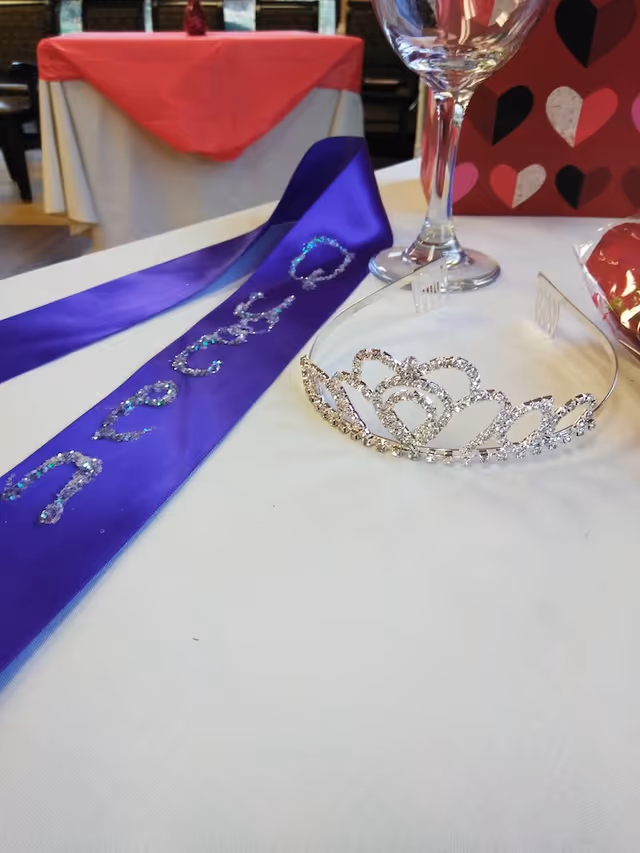 A close-up view of a table with a purple sash decorated with glittery letters, a silver tiara, a wine glass, and a red gift bag with heart patterns in the background. The table is covered with a white tablecloth and there is another table with a red tablecloth in the background.