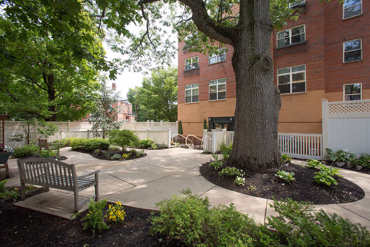 Outdoor courtyard area with a large tree surrounded by landscaped flower beds, benches, and a paved walkway. The courtyard is enclosed by a white fence and adjacent to a multi-story brick building.