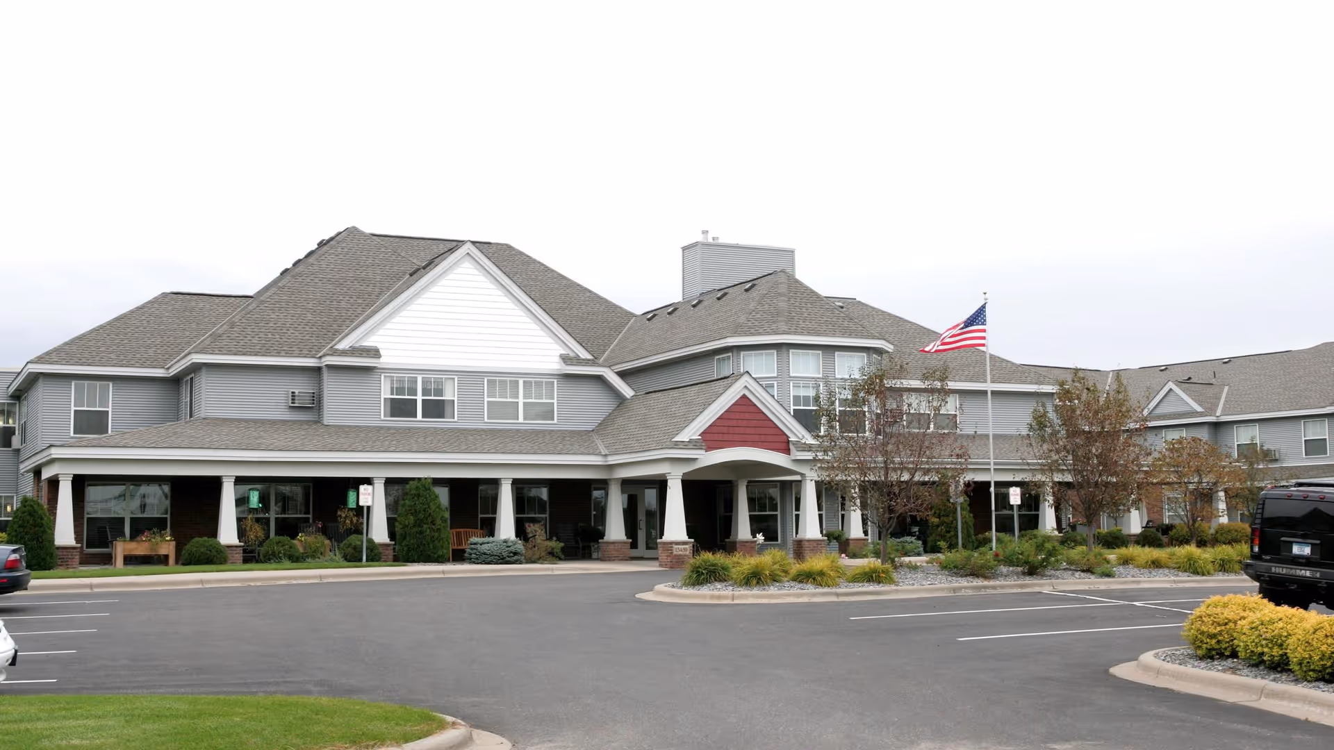 Exterior view of a senior living facility building with gray siding and a peaked roof. There is a covered entrance supported by white columns, landscaping with bushes and small trees, an American flag on a flagpole, and a parking lot in front with several parked vehicles.