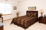 A bedroom with a large window letting in natural light, a bed with brown striped bedding, a wooden headboard, two matching nightstands with lamps, a framed picture above the bed, and a vase with dried plants near the window.