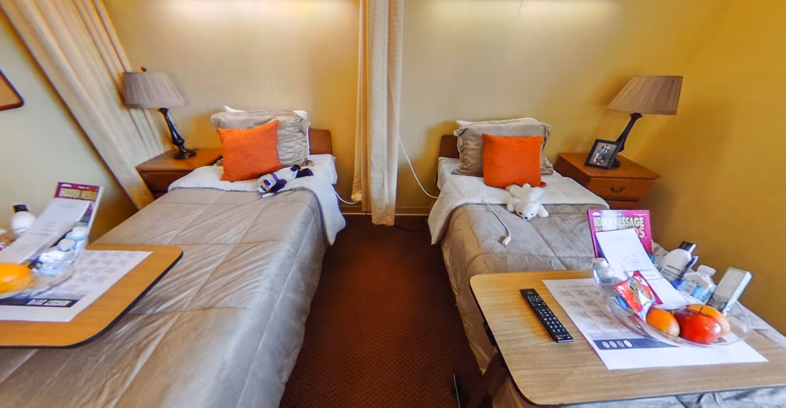 Small shared bedroom with two twin beds, orange accent pillows, bedside tables and over-bed trays with personal items.