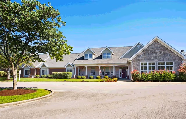 Front exterior view of a single-story senior living facility building with a stone and siding facade, multiple windows, a covered entrance with columns, landscaping including bushes and flowers, a tree on the left, and a clear blue sky.