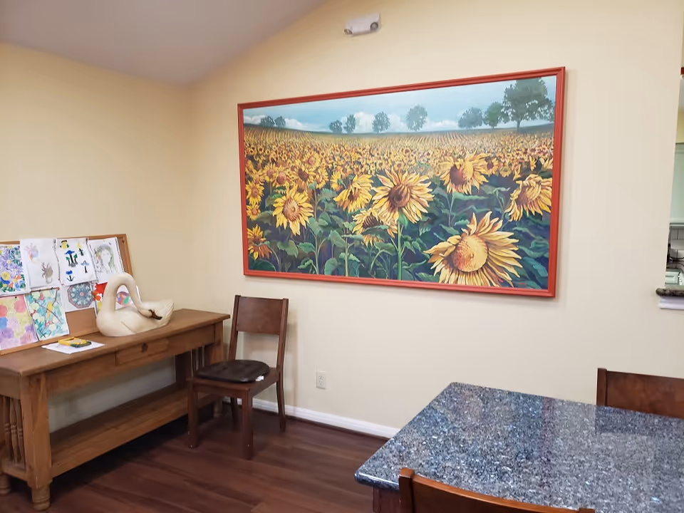 Interior common area with a large framed sunflower painting above a wooden side table and chairs and a granite-topped table in the foreground.