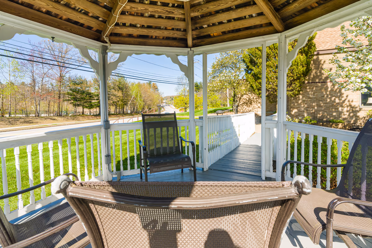 View from inside a wooden gazebo with a shingled roof, white railings, and several brown mesh chairs. The gazebo overlooks a green lawn, a sidewalk with a ramp, and a road with trees and buildings in the background under a clear blue sky.