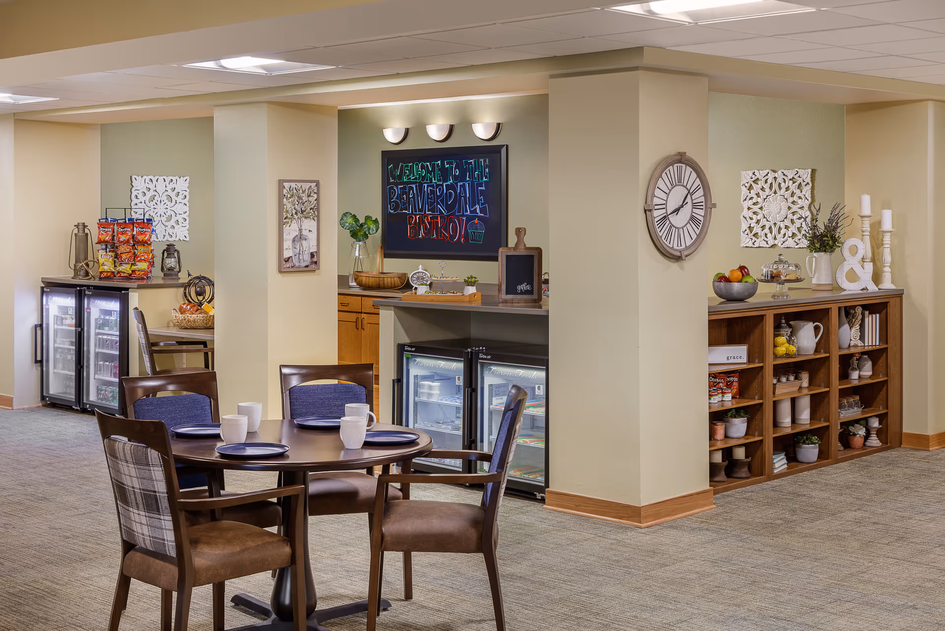 A cozy dining area in a senior living facility with a round wooden table set with four chairs, each with a plate and mug. In the background, there are two small refrigerators stocked with beverages and snacks, a chalkboard sign that reads 'Welcome to the Beaverdale Bistro!', decorative wall art, a large clock, and a wooden shelving unit with various decorative items and fruit bowls.
