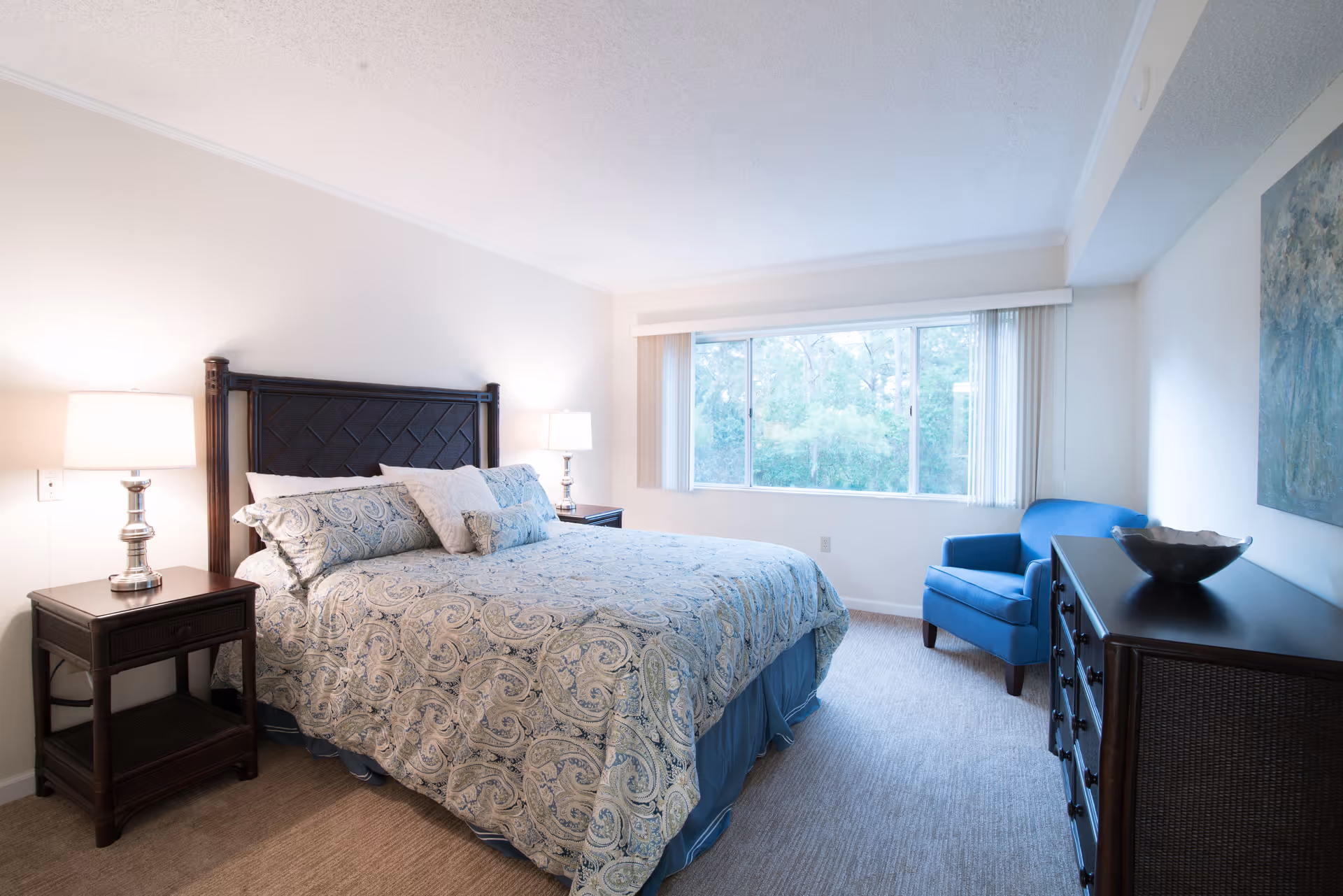 A cozy bedroom featuring a large bed with decorative bedding, two bedside lamps, and a window with a view of greenery. A blue chair and a dresser are also visible in the room.