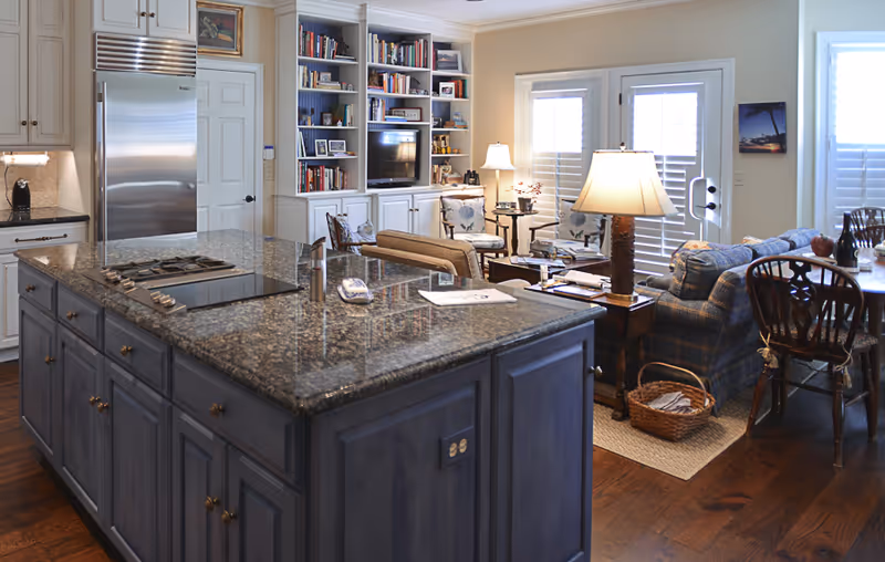 Open-plan kitchen with a large granite island in the foreground and a cozy living area with bookshelves and seating in the background.