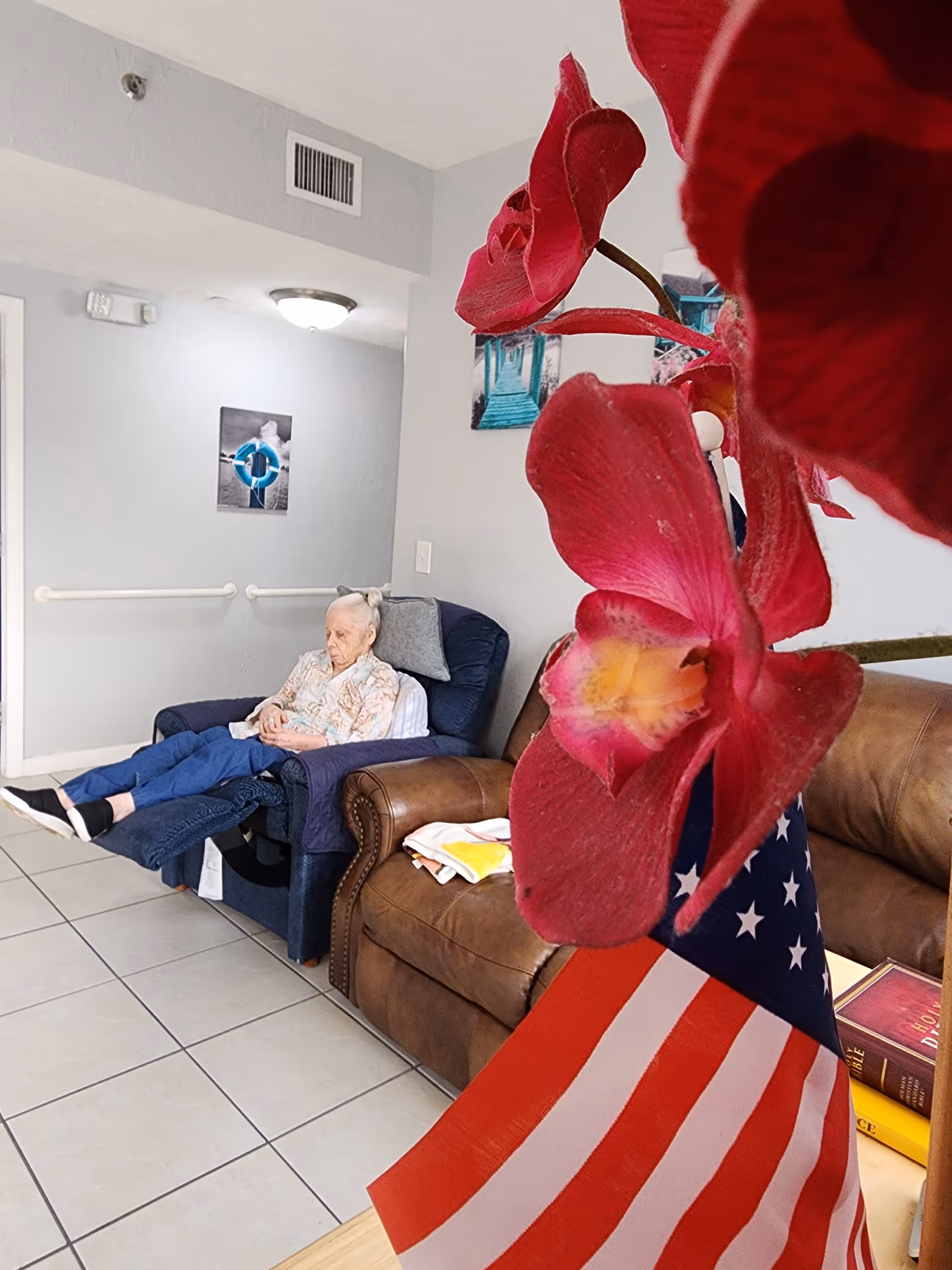 An elderly woman is sitting in a recliner chair in a living room area with tiled floors. In the foreground, there is a close-up of a red orchid flower and a small American flag. The room has a brown leather couch, a wall with a handrail, and framed pictures hanging on it.