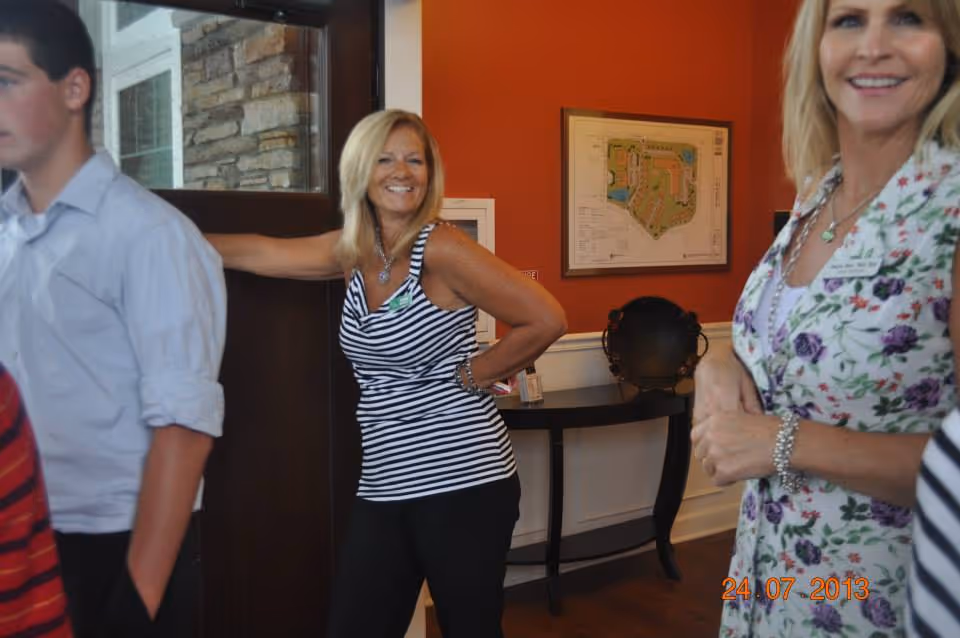 A smiling woman in a black and white striped sleeveless top stands with her hand on her hip near a door inside a room with orange walls. Another woman in a floral dress is partially visible on the right, and a young man in a light blue shirt is partially visible on the left. A framed map or floor plan hangs on the wall behind them above a small table with decorative items.