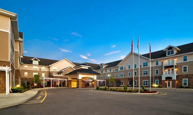 Exterior view of Pinebrook of Milford by StoryPoint Group senior living facility at dusk, showing a large three-story building with multiple windows, a covered entrance, three flagpoles with flags, and a circular driveway.