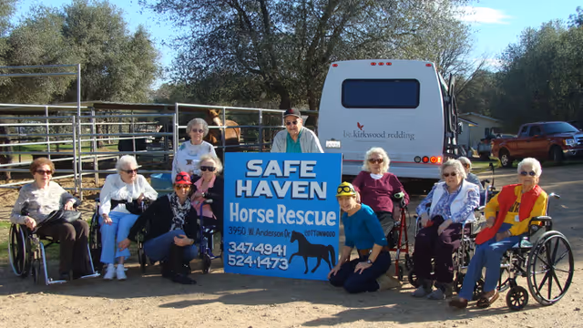 A group of elderly people, some in wheelchairs, posing outdoors around a blue sign that reads 'SAFE HAVEN Horse Rescue' with contact information. Behind them is a white vehicle and some trees.