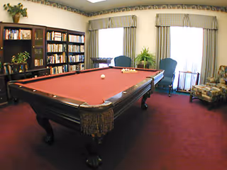 Indoor common room with a red-felt pool table, bookshelves, armchairs, and large windows with draped curtains.