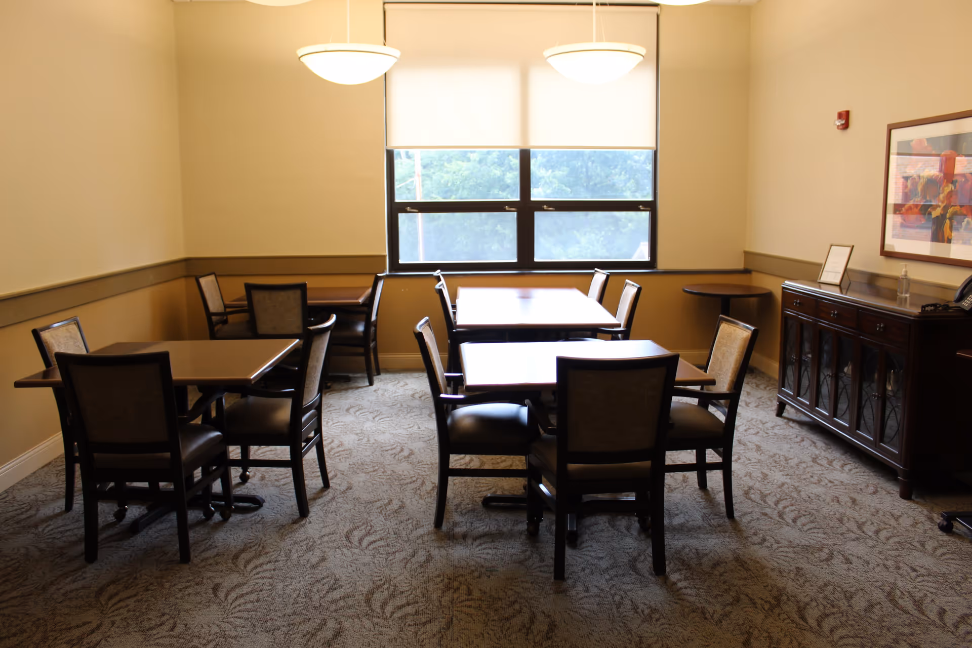 A small dining room with several wooden tables and chairs arranged neatly on a patterned carpet. Two pendant lights hang from the ceiling, and a large window with a white roller shade lets in natural light. A wooden sideboard with glass doors and a framed artwork on the wall are visible on the right side of the room.