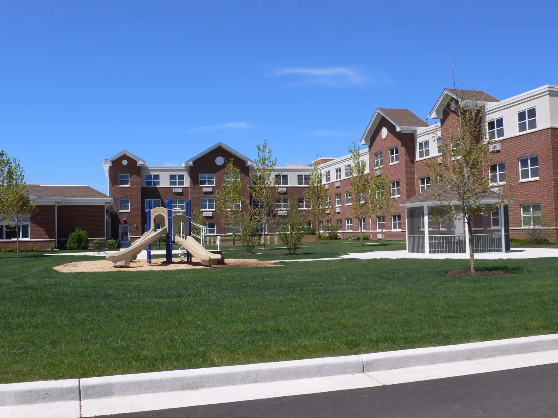 Red brick senior living building with a grassy courtyard, playground and a gazebo under a clear blue sky.