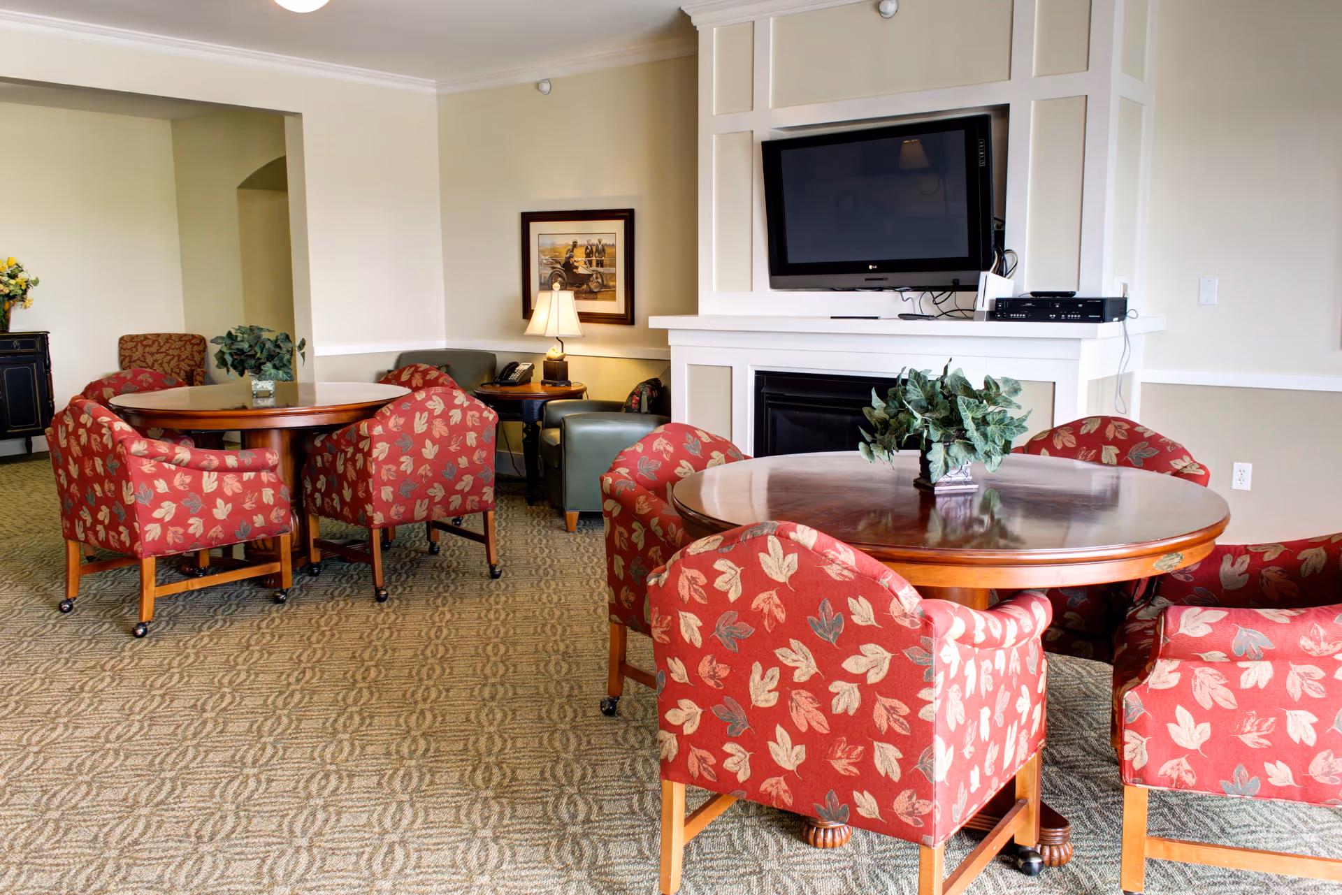 Communal lounge with round wooden tables, red patterned chairs, a wall-mounted TV above a fireplace, and a potted plant centerpiece.