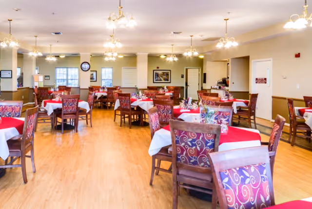 A spacious dining room with multiple tables covered in white and red tablecloths, surrounded by wooden chairs with patterned upholstery. The room has wooden flooring, beige walls, several windows, and multiple ceiling light fixtures providing warm lighting.