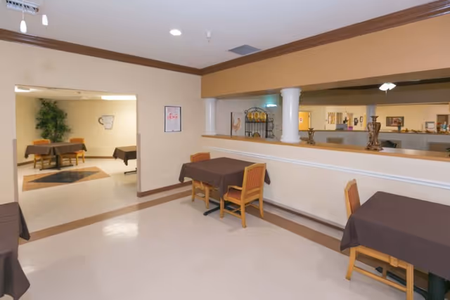 Interior view of a dining area in a senior living facility with several tables covered with brown tablecloths and wooden chairs. The room has beige walls with a brown trim and a large open window-like cutout in the wall showing another room with more tables and chairs. There are decorative items on the ledge of the cutout and framed pictures on the walls.