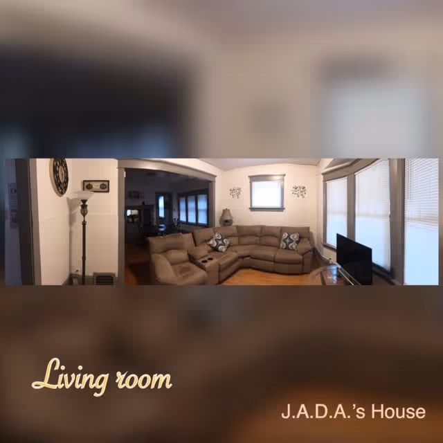 A cozy living room with a large beige sectional sofa adorned with patterned pillows. There is a floor lamp and a wall clock on the left side. A small TV is placed on a glass stand near the windows with blinds. The walls are light-colored, and there is a small window with decorative wall art above the sofa.