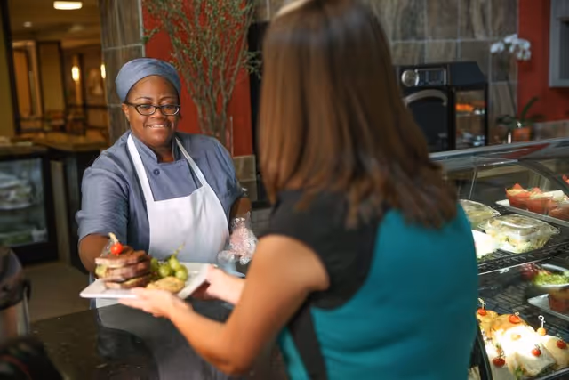 A woman wearing a gray uniform and white apron hands a plate of food with grapes and a sandwich to another woman in a teal and black top at a counter with a display case of food items in the background.