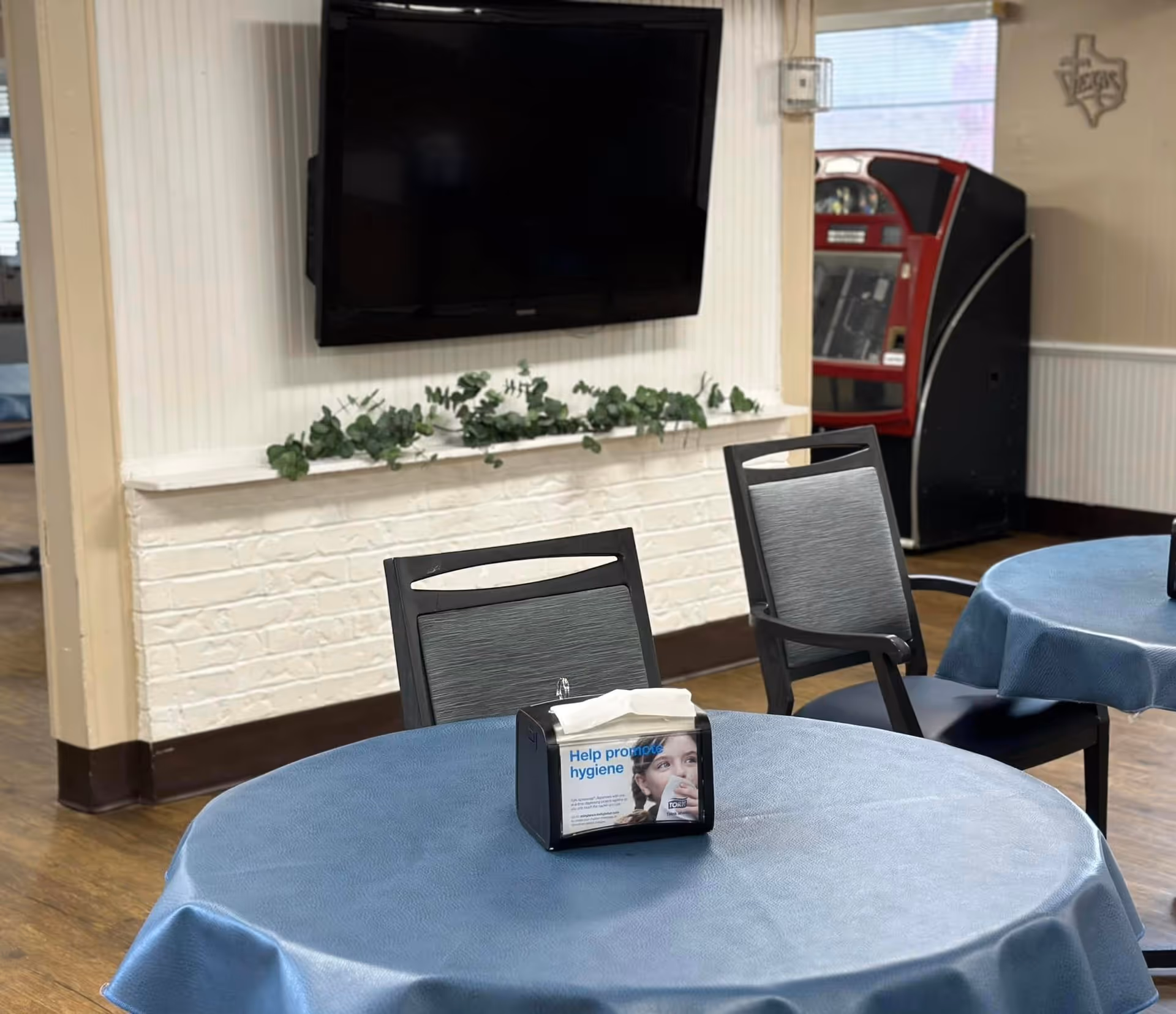 A common area with round tables covered in blue tablecloths and black chairs. A flat-screen TV is mounted on a white brick wall with a small green plant decoration below it. In the background, there is a red vending machine and a wall decoration shaped like the state of Texas.