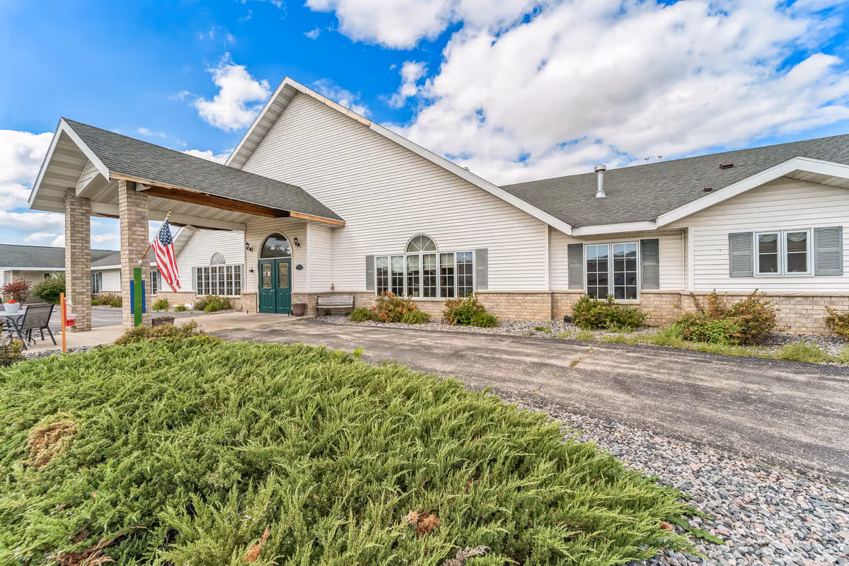 Exterior view of a single-story senior living facility building with white siding and a gray roof under a partly cloudy sky. The entrance features a covered porch with an American flag hanging, green double doors, and surrounding landscaping with bushes and a paved driveway.
