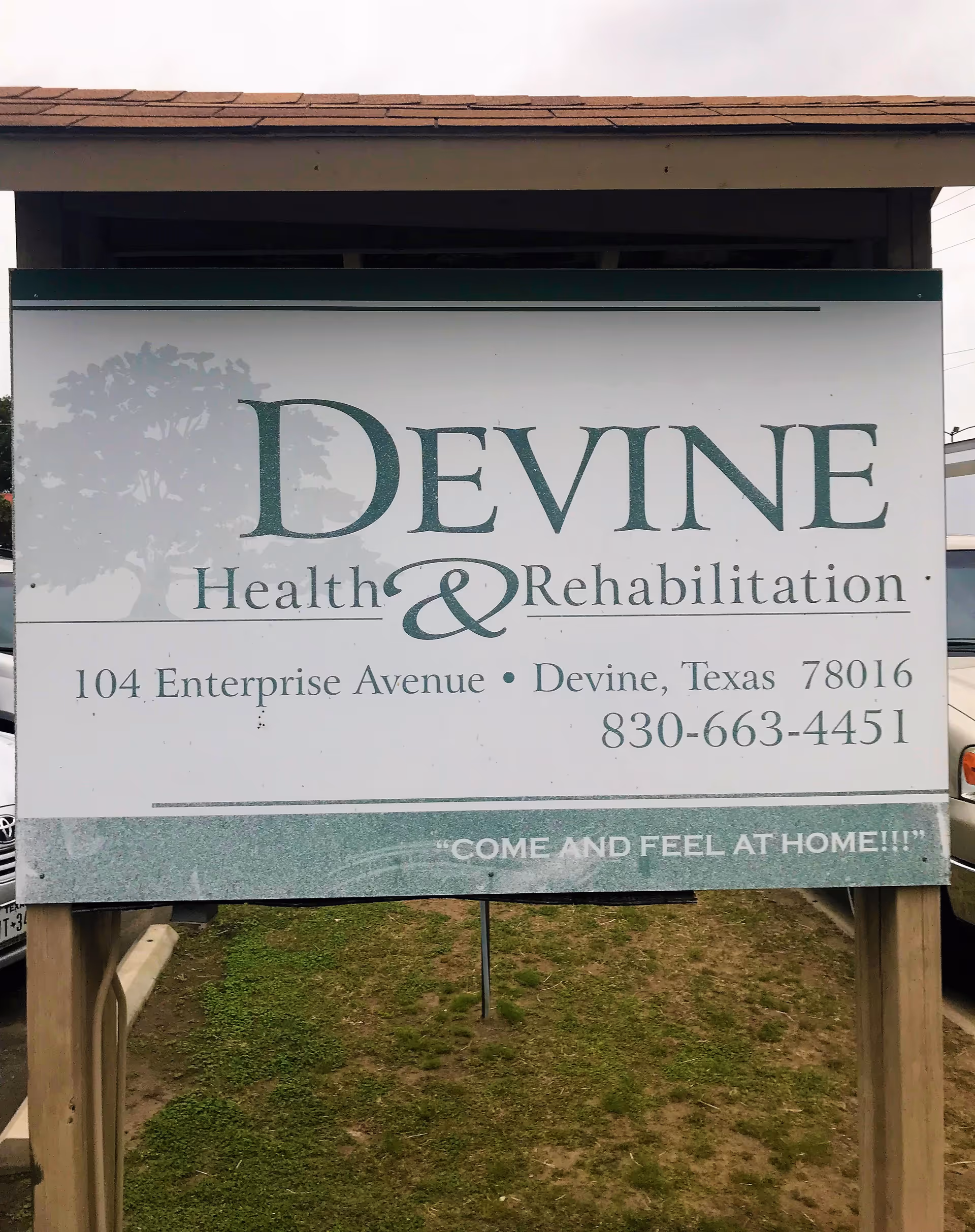 A large outdoor sign for Devine Health & Rehabilitation located at 104 Enterprise Avenue, Devine, Texas 78016, with the phone number 830-663-4451 and the slogan 'COME AND FEEL AT HOME!!!'. The sign is mounted on wooden posts with grass and parked cars visible in the background.