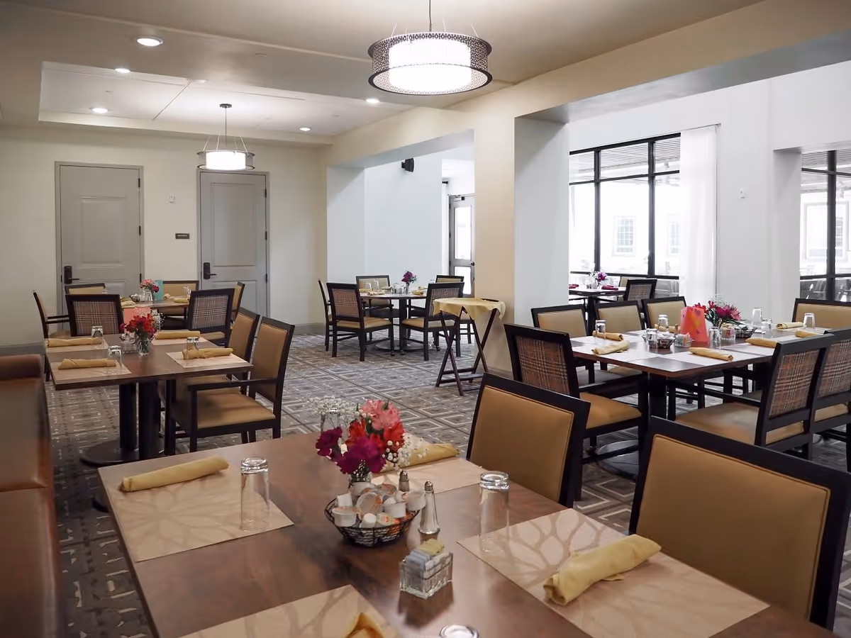 A well-lit dining room with multiple tables set with placemats, napkins, glasses, and small flower arrangements. The room features beige walls, patterned carpet, and modern light fixtures on the ceiling. There are several chairs around each table, and large windows on one side letting in natural light.