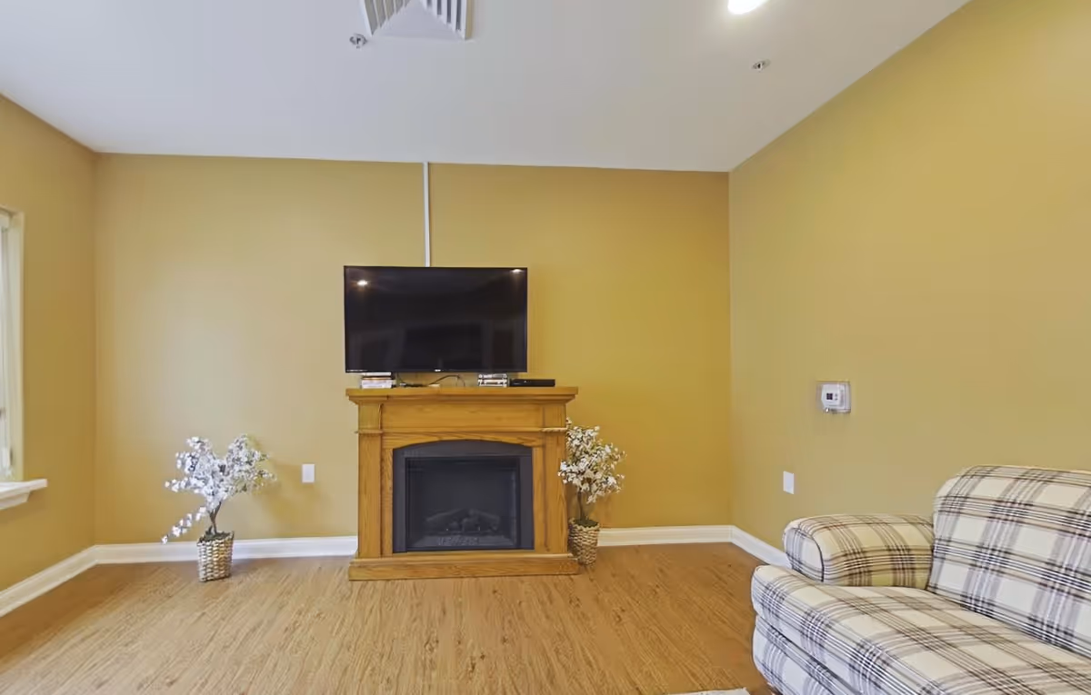 A cozy living room with a wooden floor, beige walls, a flat-screen TV mounted above a wooden electric fireplace, two potted plants with white flowers on either side of the fireplace, and a plaid upholstered armchair on the right side.