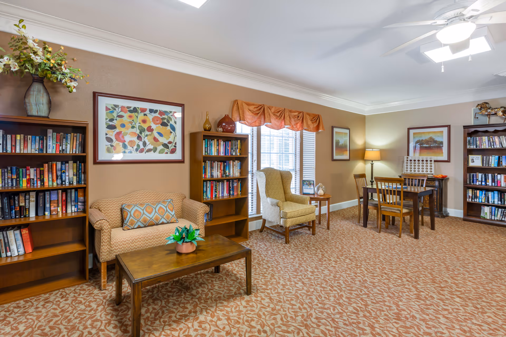 A cozy common area with beige walls and patterned carpet featuring a small sofa with a decorative pillow, a wooden coffee table with a small plant, two bookshelves filled with books, a comfortable armchair near a window with orange valance curtains, a wooden table with four chairs, a floor lamp, and framed artwork on the walls.