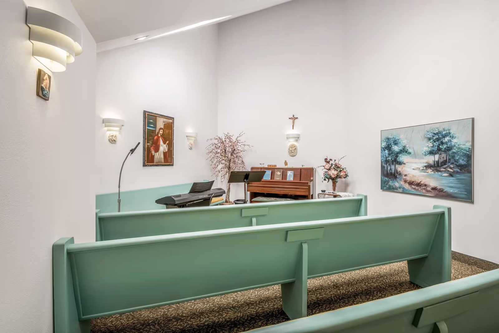 Interior of a small chapel or prayer room with light green wooden pews facing a piano and keyboard. The walls are white and decorated with religious artwork, including a painting of Jesus, a crucifix, and a landscape painting. There are wall-mounted lights and floral arrangements near the piano.