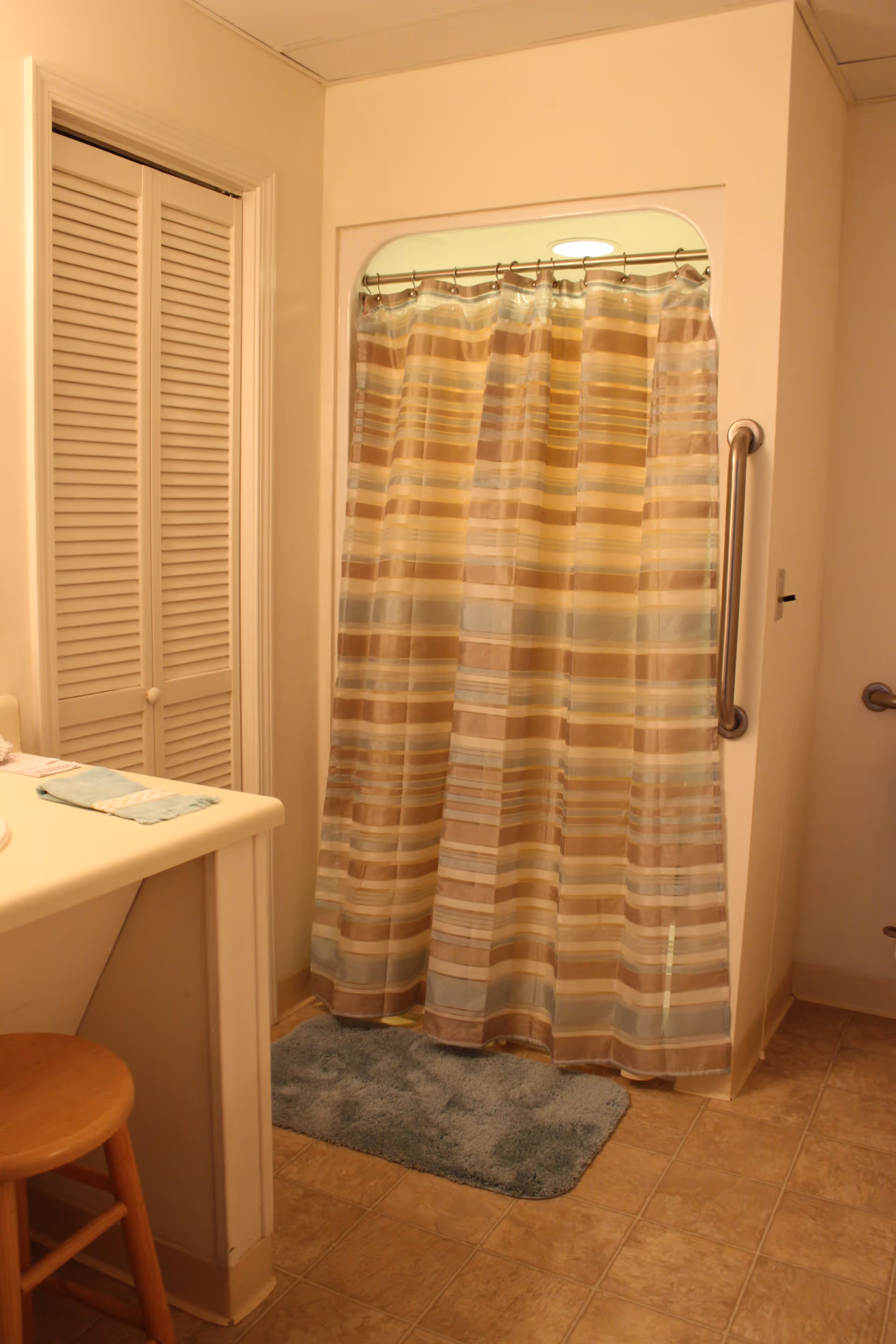 A bathroom with a shower area enclosed by a striped shower curtain in shades of beige, brown, and blue. There is a blue bath mat on the tiled floor in front of the shower. A wooden stool is placed near a white countertop with a folded towel on it. The walls are light-colored, and there is a grab bar mounted on the wall next to the shower entrance.