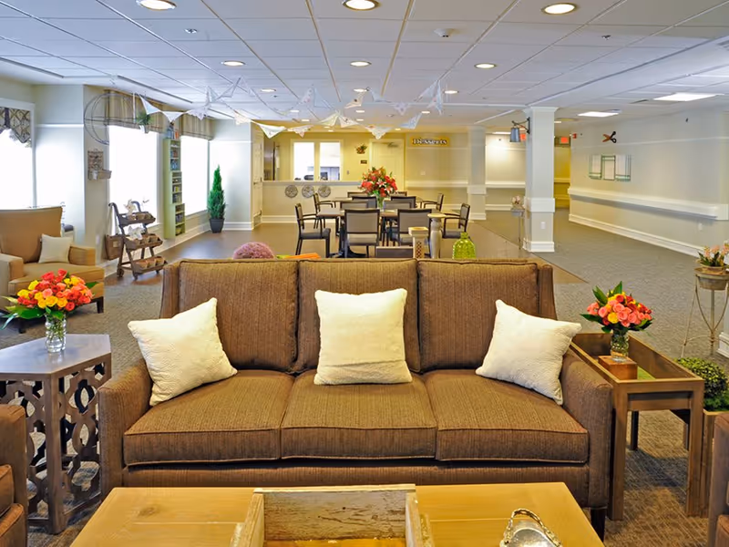 Bright communal lounge in a senior living facility with a brown sofa in the foreground, dining tables and chairs and floral arrangements in the background.
